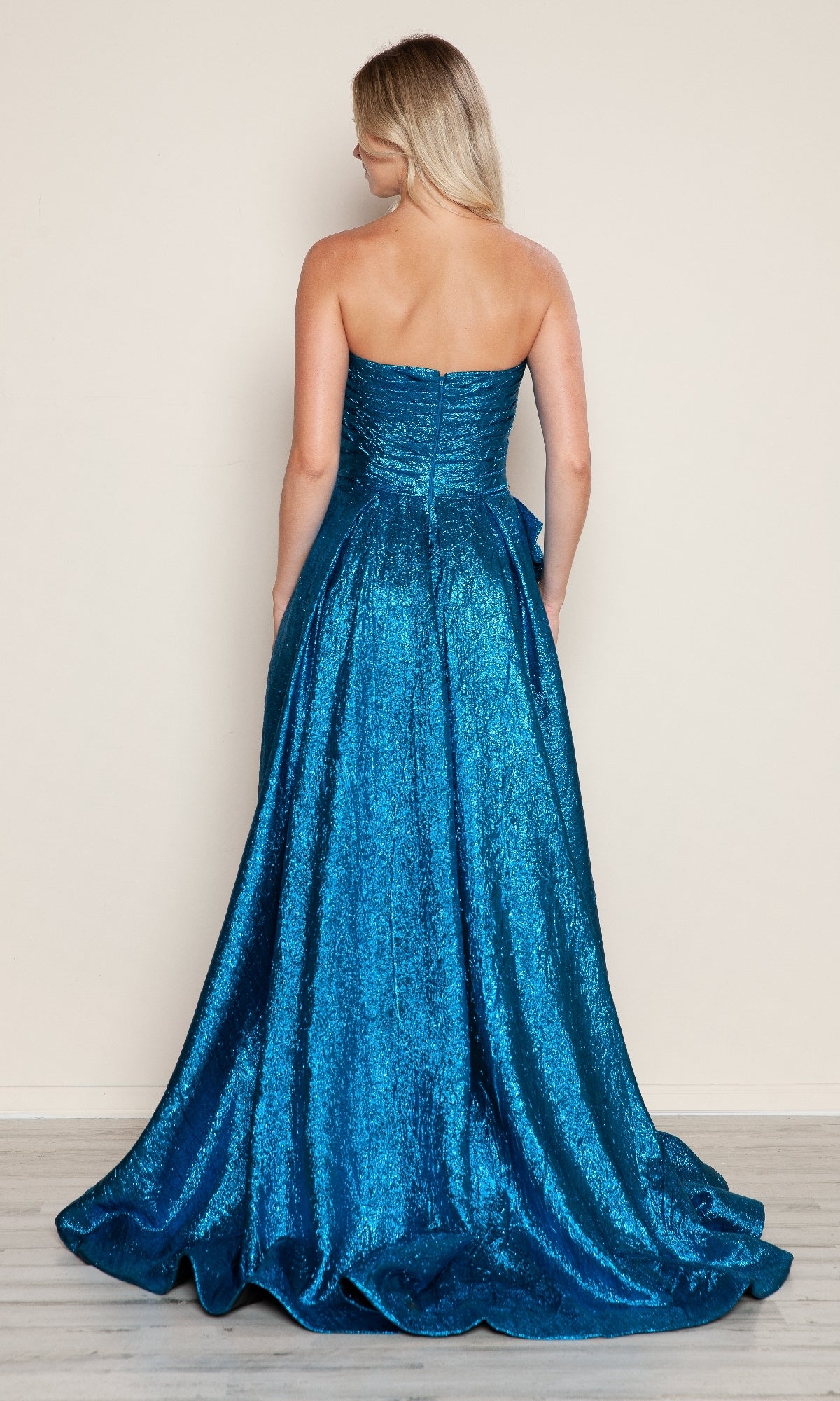 A woman with long blonde hair is shown from the back wearing the Poly USA 9720 Side-Bow Strapless Prom Gown, featuring a floor-length glitter skirt, against a plain light-colored wall.